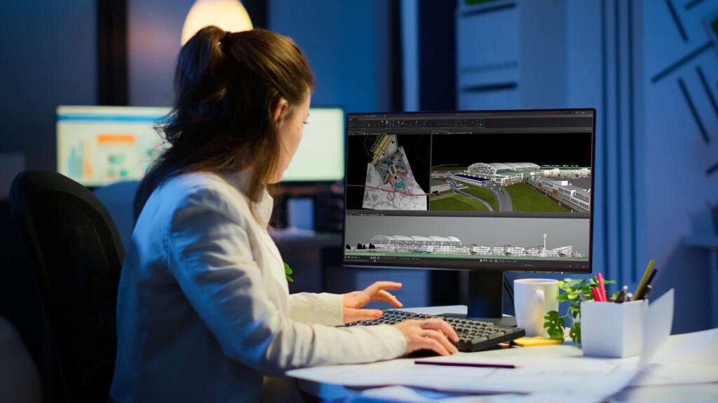 Woman at desk working on 3D architectural model on computer screen, with office equipment and lighting in the background.